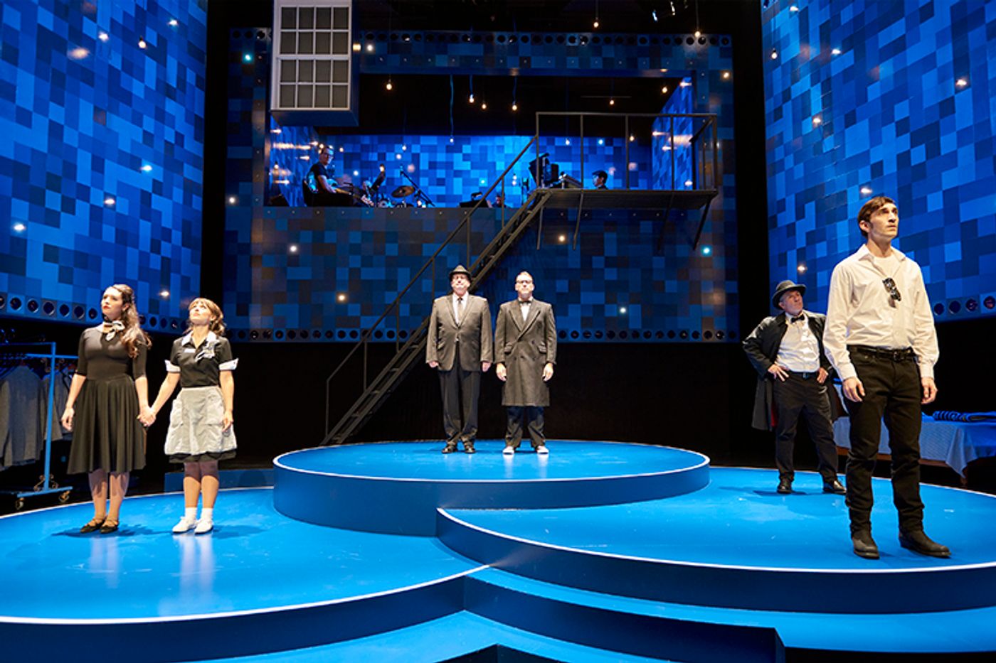 Production still of Fly by Night. Six actors stand scattered on a blue-themed stage. They gaze up at the audience. Above them hang dozens and dozens of lightbulbs, emulating stars.