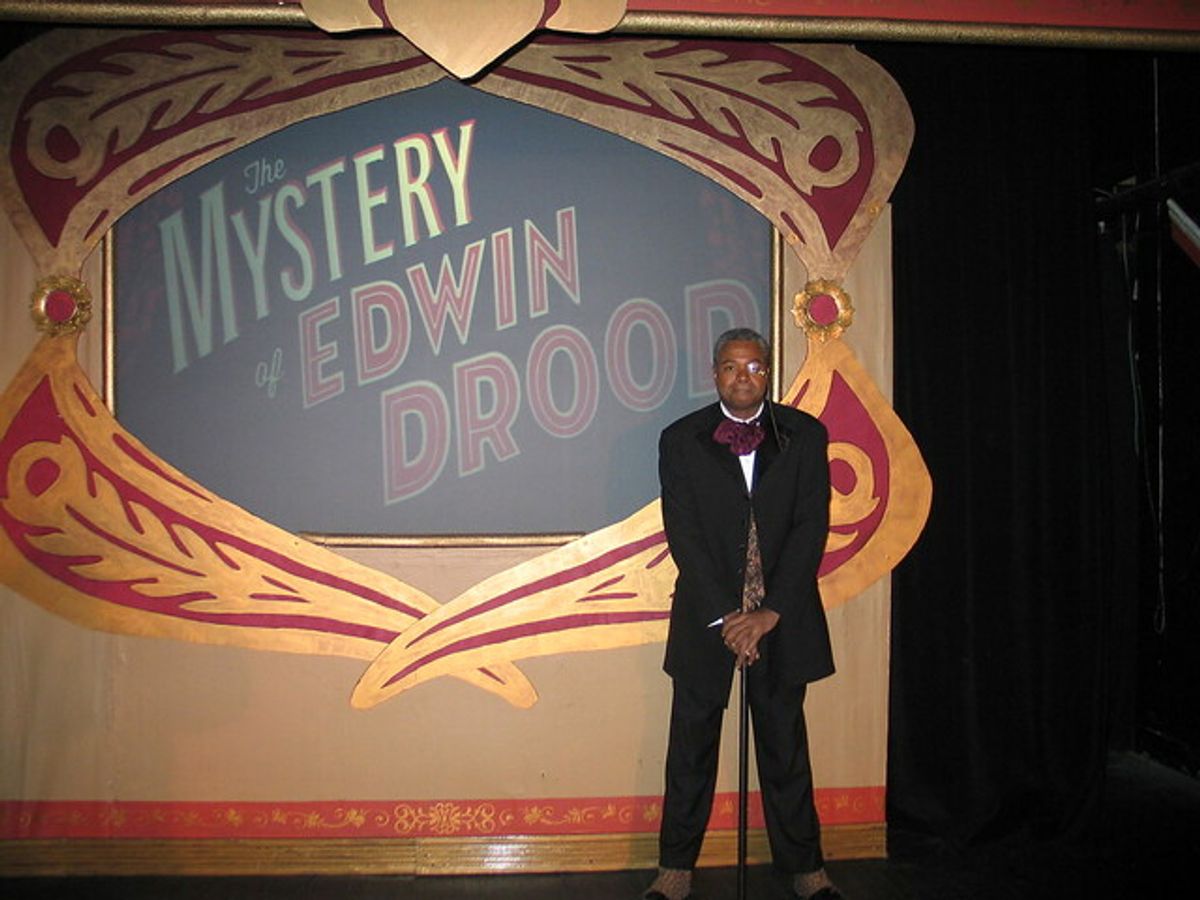 It Happens In A Place Called Cloisterham!: Darryl Maximilian Robinson as The Chairman Mr. William Cartwright and Mayor Thomas Sapsea unfolds the musical tale of an unfinished Dickens' novel in the 2018 Saint Sebastian Players of Chicago Revival of Rupert Holmes' 'The Mystery of Edwin Drood'. Photo by Set Designer Emil Zbella. It Happens In A Place Called Cloisterham!: Darryl Maximilian Robinson as The Chairman Mr. William Cartwright and Mayor Thomas Sapsea unfolds the musical tale of an unfinished Dickens' novel in the 2018 Saint Sebastian Players of Chicago Revival of Rupert Holmes' 'The Mystery of Edwin Drood'. Photo by Set Designer Emil Zbella.