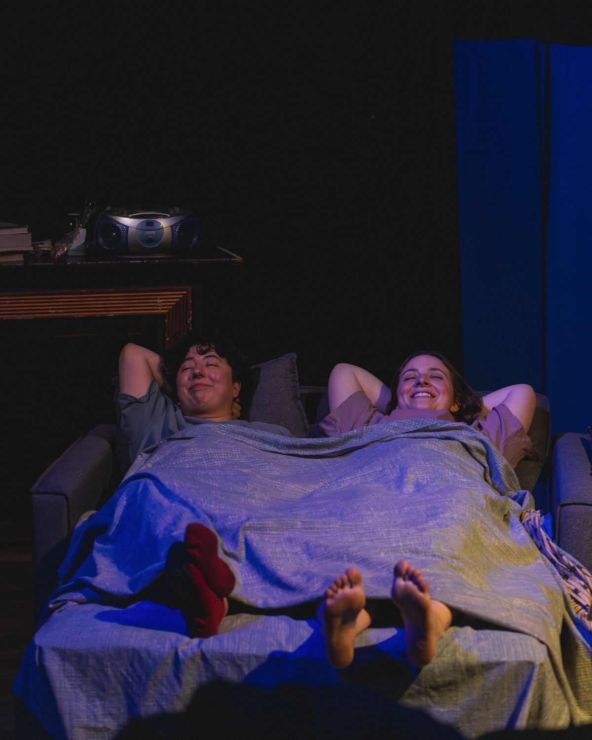Melanie Santos Cordero as Callie and Bridgette Dove as Sarah are lying down on a sofa bed in Stop Kiss. Photo by MCP Photo. Melanie Santos Cordero as Callie and Bridgette Dove as Sarah are lying down on a sofa bed in Stop Kiss. Photo by MCP Photo.