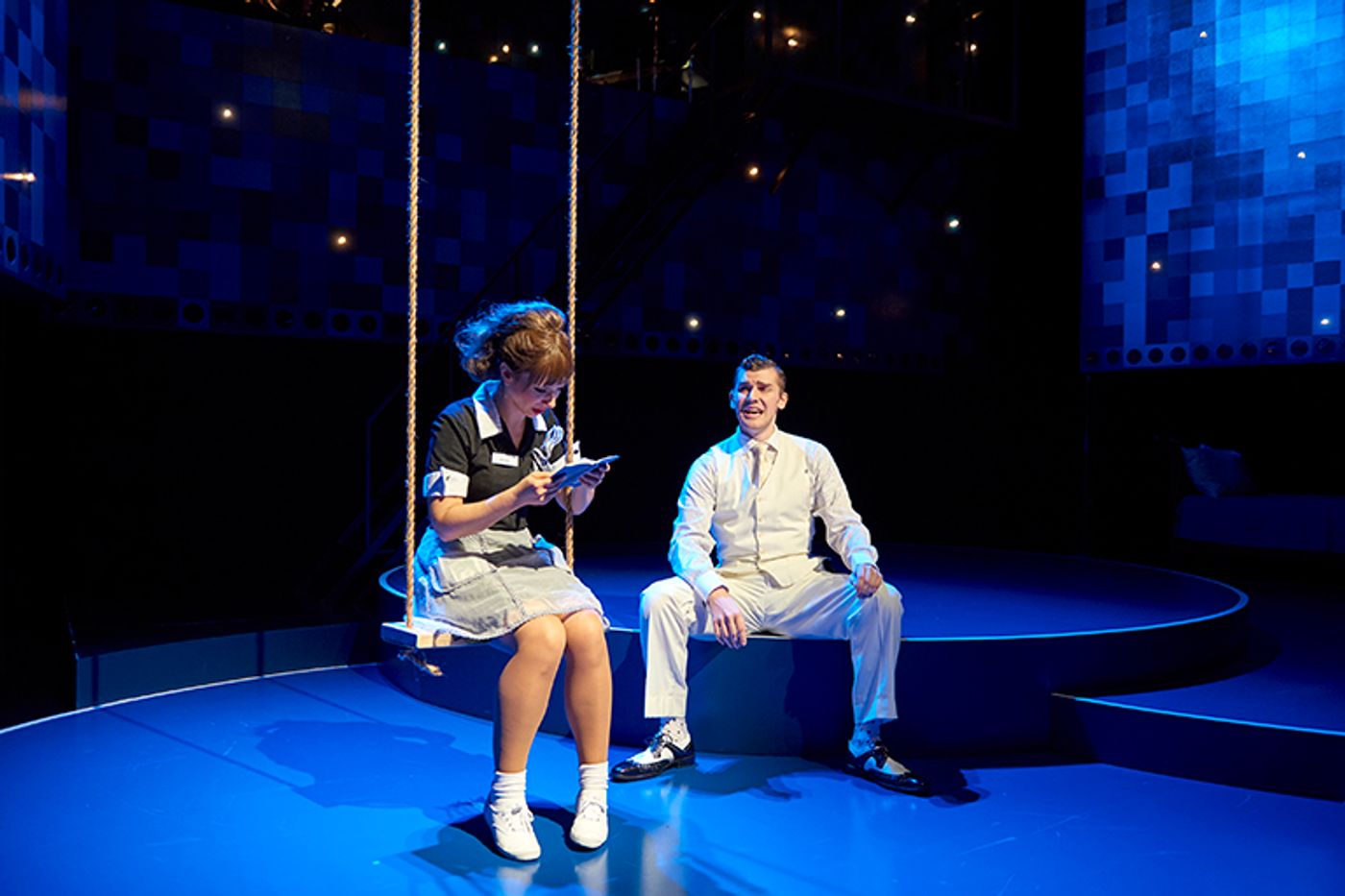 A production still from a performance of Fly by Night. A young woman with bangs and hair pulled back in a large ponytail sits on a swing with a book. A man in a white suit sits nearby.