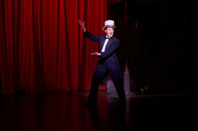 Mr. Memory’s emcee (actor, Maggie Kettering*) readies the audience for the show in  Photo