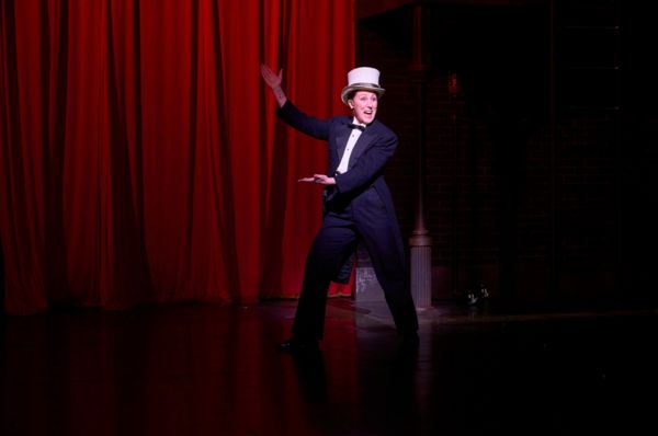 Mr. Memory’s emcee (actor, Maggie Kettering*) readies the audience for the show in  Photo