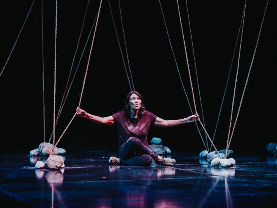 Inner Elder, Michelle Thrush, seated amongst boulders on the floor with strings leading skyward. - ©️Curtis Perry
