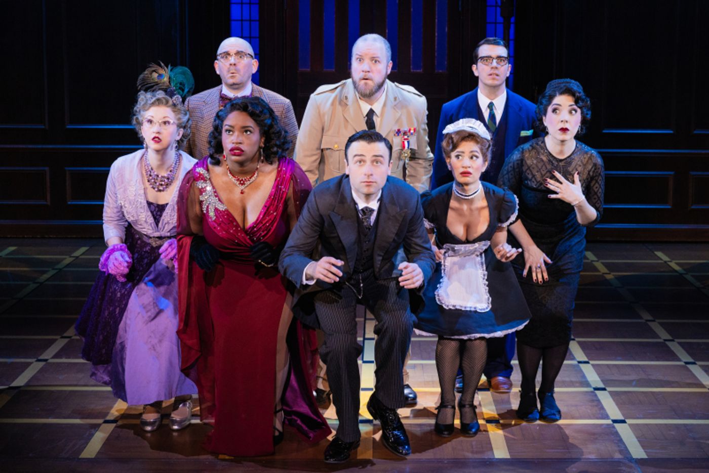 Four women and a ma n stand on stage crouched in a line with three men standing behind them as they search for something. All actors are looking to their forward off in the distance with a frightened look on their faces.