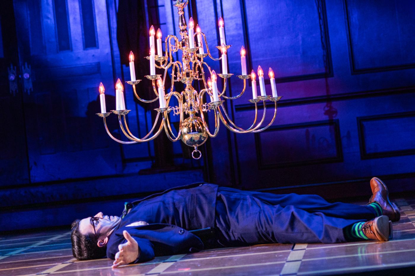 A man is lying spread out on the floor, mouth agape as a falling chandelier hangs over him. 